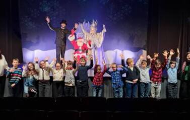 grupa uczni&oacute;w z podniesionymi rękami przed sceną w teatrze Picolo. Na scenie trzech aktor&oacute;w: na fotelu siedzi święty mikołaj, po jego prawej stronie stoi kr&oacute;lowa lodu, a po prawej w czarnym stroju i kapeluszu narrator.