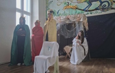 Uczniowie podczas przedsatwienia jasełek: W przebraniach siedzą w stajence Maryja z J&oacute;zefem, między kt&oacute;rymi stoi żłobek z dzieciątkiem. Po lewej stoi w przebraniach trzech kr&oacute;li trzymający dary.