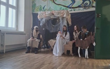 Uczniowie podczas przedsatwienia jasełek: W przebraniach w stajence siedzi J&oacute;zef i Maryja. Międzu nimi żłobek z dziciątkiem. Z prawej dwaj pasterze przyklękają i trzymają tekturowe owieczkę i osiołka