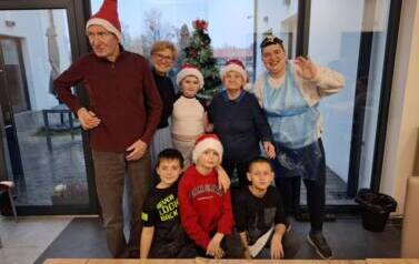 Grupa uczni&oacute;w w czapkach Mikołaja  pozuje do zdjęcia z seniorami 