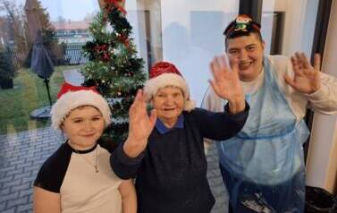 Dw&oacute;ch chłopc&oacute;w w czapkach Mikołaj a pozują do zdjęcia z uniesionymi rękami, z seniorką stoljącą po prawej stronie