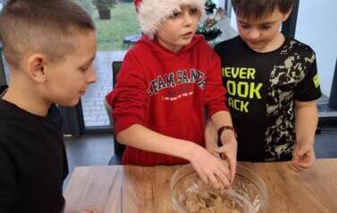 Trzech chłopc&oacute;w stoi przy stole. W środku chłopiec w przebraniu Mikołaja ugniata ciasto w misce