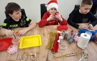 Trzech chłopc&oacute;w - w środku chłopiec w stroju Mikołaja siedzą przy stole i wykonują dekoracje świateczne