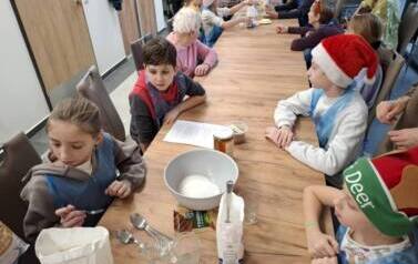 Grupa dzieci i senior&oacute;w siedząca przy stole podczas spotkania świątecznego, robią ciasteczka piernikowe