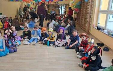 Duża grupa dzieci w przebraniach za r&oacute;żne postacie podczas balu karnawałowego siedzi w kole w sali gimnastycznej