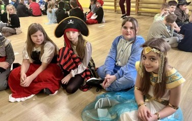 Duża grupa dzieci w przebraniach za r&oacute;żne postacie podczas balu karnawałowegosiedzi w kole w sali gimnastycznej