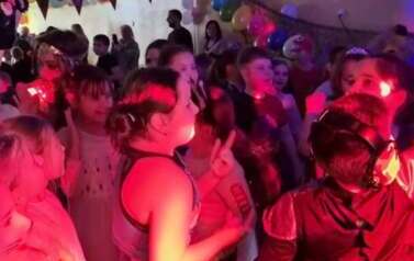 Duża grupa dzieci i nauczyciele w przebraniach za r&oacute;żne postacie podczas balu karnawałowego tańczy w sali gimnastycznej przy czerwonym tle światła