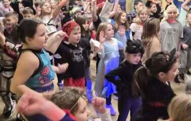 Duża grupa dzieci i nauczyciele w przebraniach za r&oacute;żne postacie podczas balu karnawałowego tańczy w sali gimnastycznej