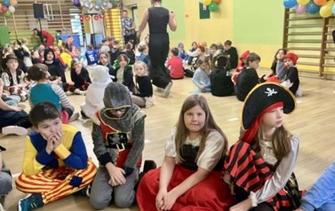 Duża grupa dzieci w przebraniach za r&oacute;żne postacie podczas balu karnawałowego siedzi w kole w sali gimnastycznej. Na pierwszym planie dziewczynka w przebraniu pirata