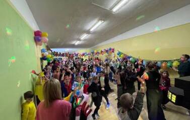 Duża grupa dzieci i nauczyciele w przebraniach za r&oacute;żne postacie podczas balu karnawałowego tańczy w sali gimnastycznej