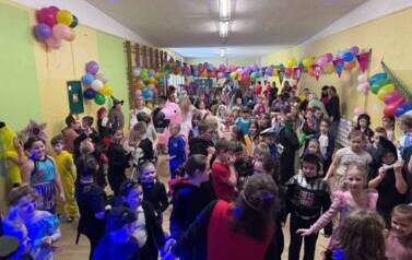 Duża grupa dzieci i nauczyciele w przebraniach za r&oacute;żne postacie podczas balu karnawałowego tańczy w sali gimnastycznej