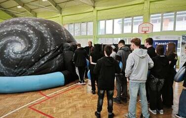 Grupa dzieci stoi gęsiego przed wejściem do kopuły mobilnego planetarium, w sali gimnastycznej.  Pan w czarnej koszulce i szarych spodniach stoi przy wejściu do kopuły mobilnego planetarium z Centrum Nauki Kopernik. 