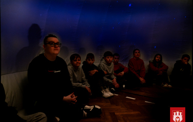 Grupa dzieci siedzi w środku kopuły mobilnego planetarium z Centrum Nauki Kopernik. Ogląda gwieździste niebo na kopule.