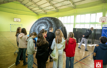 Grupa dzieci stoi gęsiego przed wejściem do kopuły mobilnego planetriaum, w sali gimnastycznej.  Pan w czarnej koszulce i szarych spodniach stoi przy wejściu do kopuły mobilnego planetarium z Centrum Nauki Kopernik. Po prawej stronie banery szkoły i Centr
