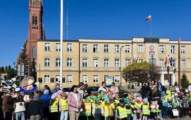 Duża grupa dzieci z elementami przebrania za jeża, stoi na placu przed Urzędem Miasta z Papierowym modelem jeża..