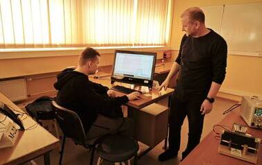 Uczeń siedzi przy stoliku przed komputerem w sali dydaktycznej Instytutu Fizyki Uniwersytetu Ł&oacute;dzkiego. Obok stoi prowadzący zajęcia