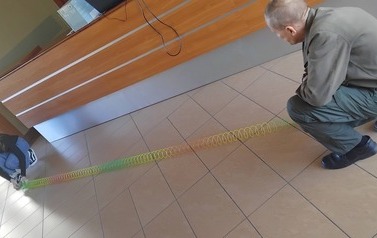 Na korytarzu Instytutu Fizyki prowadzący pracownik Uniwersyetu wykonuje z uczniem doświadczenie z rozciągnięta sprężyną 