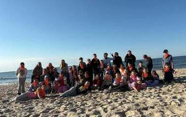 Duża grupa uczni&oacute;w pozuje do zdjęcia na nadmorkiej plaży. W tle morze.