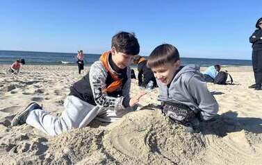 Dw&oacute;ch uczni&oacute;w w pomarańczowych chustkach na szyi buduje z piasku na nadmorskiej plaży
