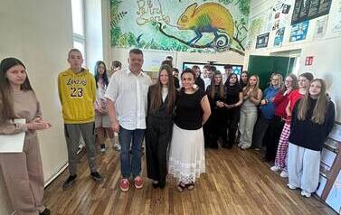 Duża grupa uczni&oacute;w wraz z dwoma opiekunami stoi na tle ektranu monitora z wyświetlonym tytułem prezentowanego projektu. Obok monitora duża makieta ze środkami uzależniającymi i ich opisem.