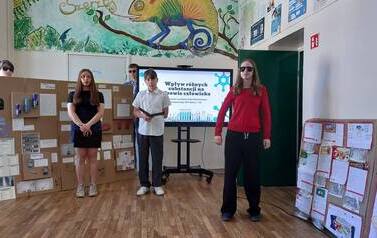 Troje uczni&oacute;w stoi na tle ektranu monitora z wyświetlonym tytułem prezentowanego projektu. Obok monitora duża makieta ze środkami uzależniającymi i ich opisem. Dziewczynka po prawej stronie w koszylce z dekoracją uszkodzonych płuc trzyma w ręku papierowy 
