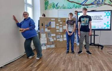 Troje uczni&oacute;w stoi na tle ektranu monitora z wyświetlonym tytułem prezentowanego projektu. Obok monitora duża makieta ze środkami uzależniającymi i ich opisem. Dziewczynka w przebraniu za ciężarną kobietę opiera się ręką o ścianie w celu utrzymaniu r&oacute;wnow