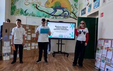 Troje uczni&oacute;w stoi na tle ektranu monitora z wyświetlonym tytułem prezentowanego projektu. Obok monitora duża makieta ze środkami uzależniającymi i ich opisem. 