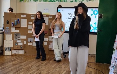 Troje uczni&oacute;w stoi na tle ektranu monitora z wyświetlonym tytułem prezentowanego projektu. Obok monitora duża makieta ze środkami uzależniającymi i ich opisem. Dziewczynka w przebraniu za osobę z problemami ukrywającą twarz pod czarnym kapturem