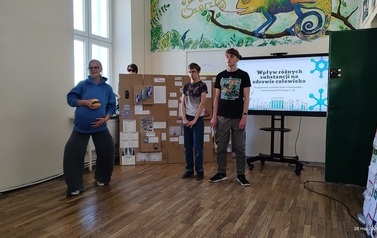 Troje uczni&oacute;w stoi na tle ektranu monitora z wyświetlonym tytułem prezentowanego projektu. Obok monitora duża makieta ze środkami uzależniającymi i ich opisem. Dziewczynka w przebraniu za ciężarną kobietę trzyma w ręku hamburgera 