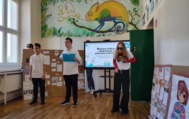 Troje uczni&oacute;w stoi na tle ektranu monitora z wyświetlonym tytułem prezentowanego projektu. Obok monitora duża makieta ze środkami uzależniającymi i ich opisem. 