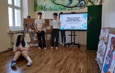 Dwoje uczni&oacute;w stoi na tle ektranu monitora z wyświetlonym tytułem prezentowanego projektu. Obok monitora duża makieta ze środkami uzależniającymi i ich opisem. Na podłodze przed stojącymi przebrana za osobę nietrzeźwą siedzi dziweczynka.