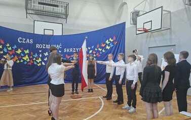 Grupa uczni&oacute;w &oacute;smych klas składa ślubowanie na sali gimnastycznej. Stoją w galowych strojach po dw&oacute;ch stronach flagi państwoej z podniesionymi w jej kierunku prawymi rękami. W tle granatowa dekoracja: na materiale kolorowe motyle