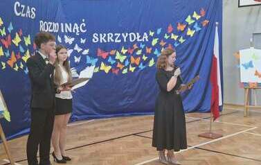 Przewodnicząca Rady Rodzic&oacute;w przemawia do uczni&oacute;w podczas zakończenia roku szkolnego w sali gimnastycznej. W tle granatowa dekoracja z kolorowymi motylami i napisem czas rozwinąc skrzydła. Po lewej stoja w strojach galowych uczniowie prezenterzy.