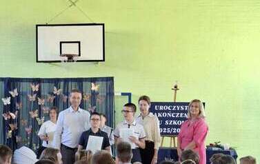 Dyrektor szkoły i wychowawca wręczają świadectwa z wyr&oacute;żnieniem i nagrody grupie uczni&oacute;w podczas zakończenia roku szkolnego w sali gimnastycznej. W tle granatowa dekoracja z kolorowymi motylami i napisem zakończenie roku szkolnego. 