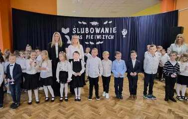 Duża grupa uczni&oacute;w klas 1 w sali gimnastycznej podczas występu i ślubowania. w tle na czarnym materiale napis Slubowanie klas pierwszych
