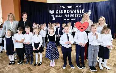 Duża grupa uczni&oacute;w klas 1 w sali gimnastycznej podczas występu i ślubowania. w tle na czarnym materiale napis Slubowanie klas pierwszych