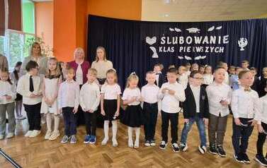 Duża grupa uczni&oacute;w klas 1 w sali gimnastycznej podczas występu i ślubowania. w tle na czarnym materiale napis Slubowanie klas pierwszych