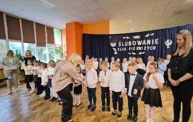 Duża grupa uczni&oacute;w klas 1 w sali gimnastycznej podczas występu i ślubowania. w tle na czarnym materiale napis Slubowanie klas pierwszych