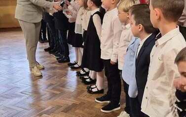 Duża grupa uczni&oacute;w klas 1 w sali gimnastycznej podczas występu i ślubowania. w tle na czarnym materiale napis Slubowanie klas pierwszych