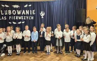 Duża grupa uczni&oacute;w klas 1 w sali gimnastycznej podczas występu i ślubowania. w tle na czarnym materiale napis Slubowanie klas pierwszych