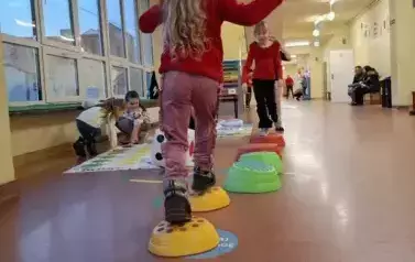 Drzwi otwrte szkoły. Na szkolnym korytarzu ułożne kolorowe klocki sensoryczne. Po klockach spacerują dwie dziewczynki w czerownych bluzkach. Jedna tyłem, druga idąca w jej kierunku przodem. Obok klock&oacute;w na macie na podłodze ułożone inne klocki sensoryczne