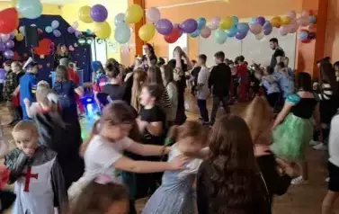 Duża grupa dzieci w r&oacute;żnych kolorowych przebraniach karnawałowych, tańczy ustawiona w węża w całej sali. W tle ubrana na czarno pani prowadząca zabawę. Dzieci bawią sie w dużej sali udekorowanej dużą ilością kolorowych balon&oacute;w zawieszonych nad ich głowami
