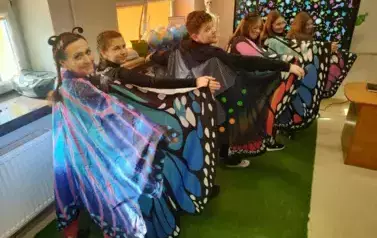 Grupa 6 os&oacute;b stojąca tyłem na tle okna z rozłożonymi pelerynami jak skrzydła motyla. Głowy stojących skierowane w prawą stronę pokazują uśmiechnięte, pomalowane kolorowo, twarze.