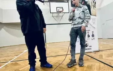 Muzyk-raper w czarnej koszulce w sali gimnastycznej, podczas występu na tle baneru z napisami RYMcerze - niezmarnuj swojego życia i zdjęć reklamujących zesp&oacute;ł. Na pierwszym planie głowy uczni&oacute;w od tyłu siedzących i oglądających spektakl. Obok muzyka stoi 