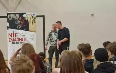 Muzyk-raper w czarnej koszulce w sali gimnastycznej, podczas występu na tle baneru z napisami RYMcerze - niezmarnuj swojego życia i zdjęć reklamujących zesp&oacute;ł. Obok muzyka stoi chłopiec w szarej bluzie i ciemnych spodniach. Na pierwszym planie głowy uczni