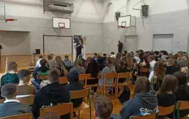 Muzyk-raper w czarnej koszulce w sali gimnastycznej, podczas występu na tle baneru z napisami RYMcerze - niezmarnuj swojego życia i zdjęć reklamujących zesp&oacute;ł. Na pierwszym planie głowy uczni&oacute;w od tyłu siedzących i oglądających spektakl.