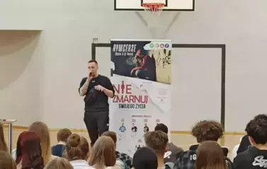 Muzyk-raper w czarnej koszulce w sali gimnastycznej, podczas występu na tle baneru z napisami RYMcerze - niezmarnuj swojego życia i zdjęć reklamujących zesp&oacute;ł. Na pierwszym planie głowy uczni&oacute;w od tyłu siedzących i oglądających spektakl.