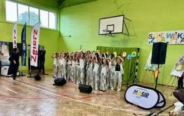 Grupa dzieci w sali gimnastycznej ubrana w białe koszulki i srebrene błyszczące spodnie podczas występu na sali gimnastycznej na imprezie Parada jeża ze Zgierza. Na ścianie napis podający tytuł imprezy.