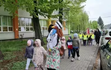 Grupa dzieci ubranych kolorowo maszerująca chodnikiem przed szkołą podczas Parady jeża ze Zgierza. Nauczycielka niesie kukłę jeż-pielędgniarka na konkurs organizowany poczas parady.