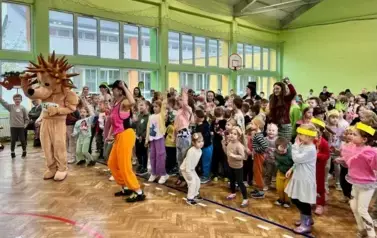 Duża grupa dzieci i opiekun&oacute;w tańczy w sali gimnastycznę podczas zakończenia imprezy Parada jeża ze Zgierza. Na czele grupy tańczy nauczycielka w pomarańczowych spodniach, czarnej bluzce i czarnej czapeczce prowadzi taniec zumbę, zachęca dzieci do zabawy.